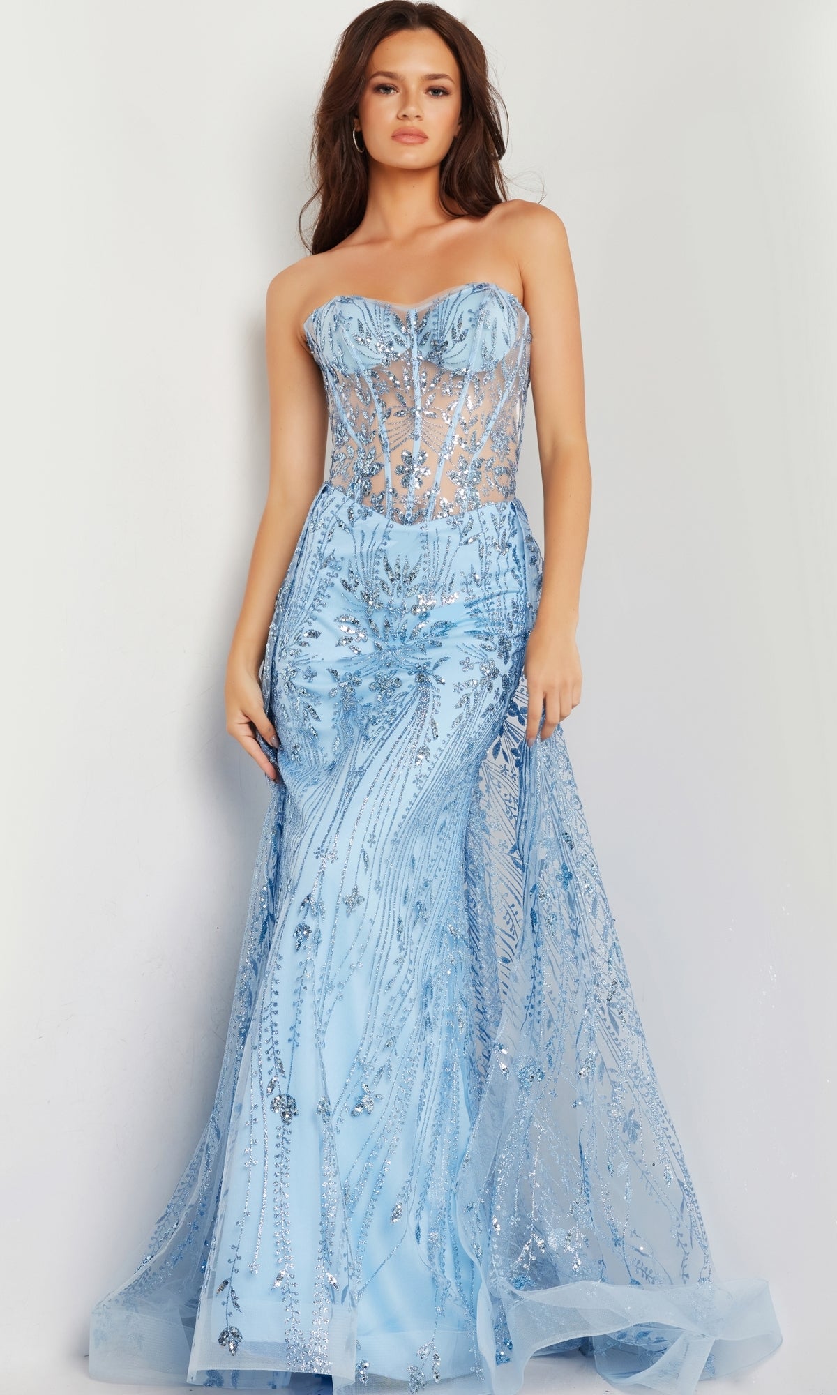 A woman models the Jovani Strapless Long Glitter-Sequin Prom Dress 26113, featuring a sheer corset bodice and a flowing mermaid skirt, standing against a plain background.